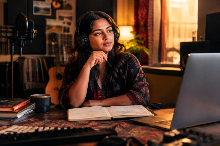 Woman in recording room, focused on songwriting with laptop and notebook.